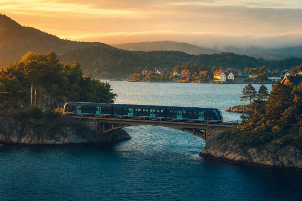 A navy-and-teal metro train crossing a low bridge over a blue strait between two green rocky islands.