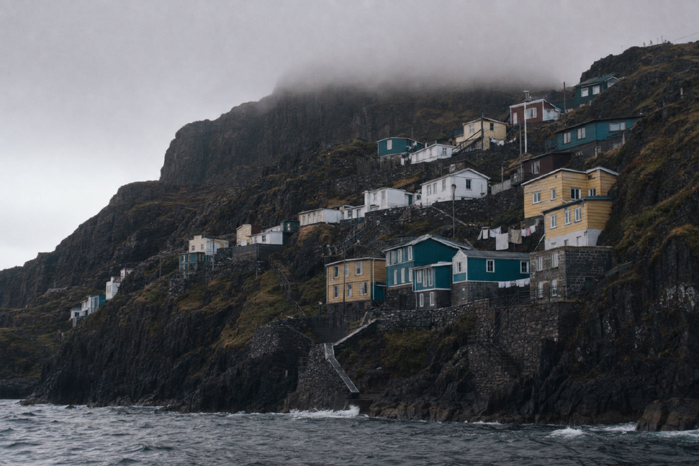 Painted timber and stone houses in stepped terraces on a steep dark sea cliff, seen from the water, with cloud sitting on the cliff top.