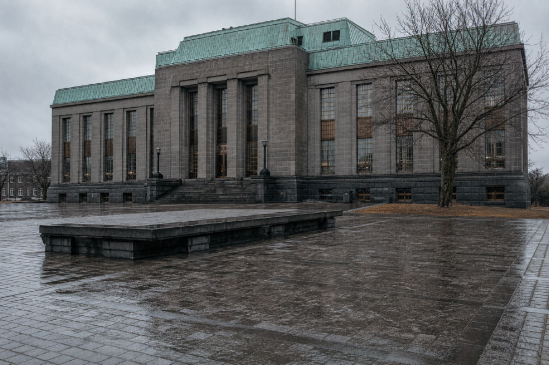 A 1930s civic building of grey basalt ashlar with a green copper roof and tall bronze windows, seen across an empty wet paved square.