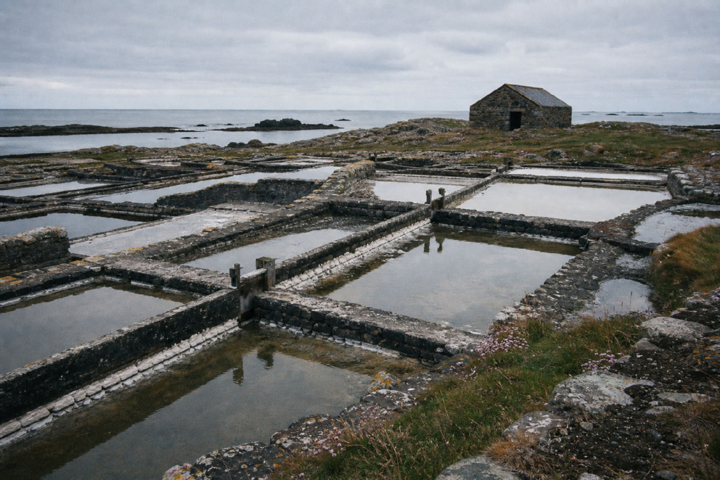 A grid of shallow salt evaporation pans divided by low stone walls on a flat island, holding still water under a grey sky, with a stone hut and the open sea beyond.