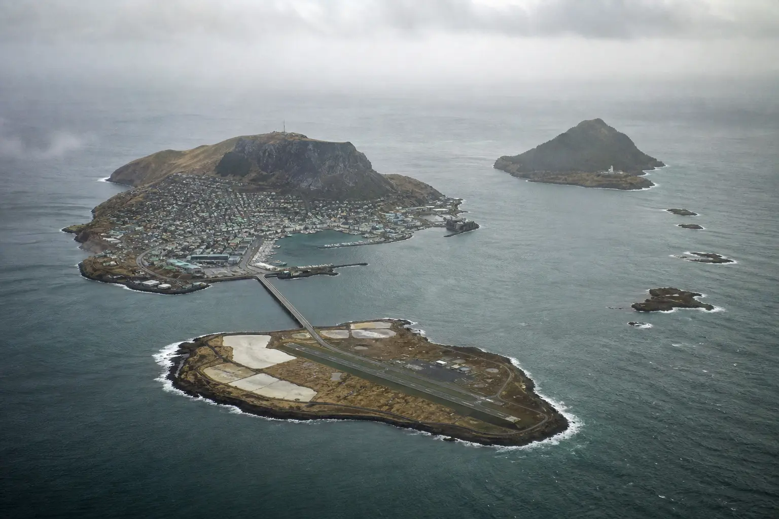 Aerial view of the Tolvaren archipelago on a clear day: the main island with the old town on its crag, the low southern island with the airport, the steep forested northern island, and three small outer isles trailing east into the sea.